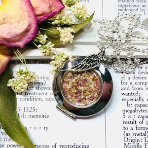 May include: A silver locket necklace with a small angel wing charm. The locket is open and filled with dried flowers and herbs. The necklace is on a white surface with text in the background.