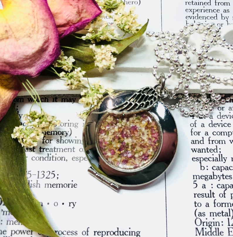 May include: A silver locket necklace with a small angel wing charm. The locket is open and filled with dried flowers and herbs. The necklace is on a white surface with text in the background.