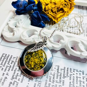 May include: A silver locket necklace with a small angel wing charm. The locket has a clear glass window that reveals a pressed flower with yellow and green petals.