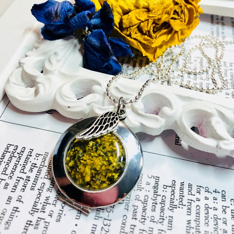 May include: A silver locket necklace with a small angel wing charm. The locket has a clear glass window that reveals a pressed flower with yellow and green petals.