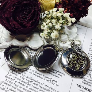 May include: A silver locket necklace with a small, round locket that opens to reveal a space for a photo or small object. The locket is attached to a silver chain with a small, silver angel wing charm. The locket is lying on a book with the word "MEMORY" printed on the page.