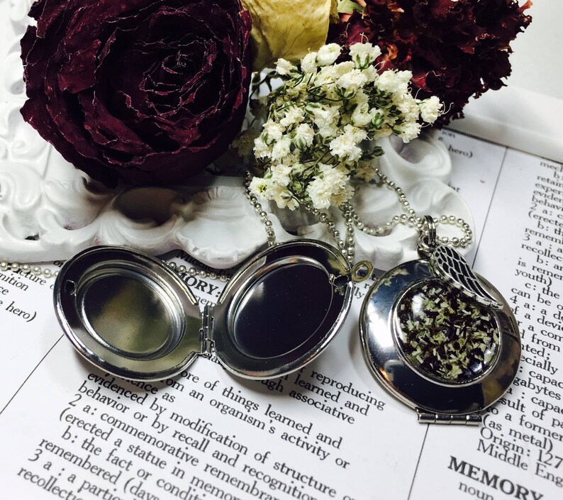 May include: A silver locket necklace with a small, round locket that opens to reveal a space for a photo or small object. The locket is attached to a silver chain with a small, silver angel wing charm. The locket is lying on a book with the word "MEMORY" printed on the page.