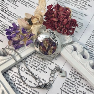 May include: A silver locket necklace with a delicate angel wing charm. The locket is open, revealing dried flowers pressed inside. The necklace is on a white surface with a book.