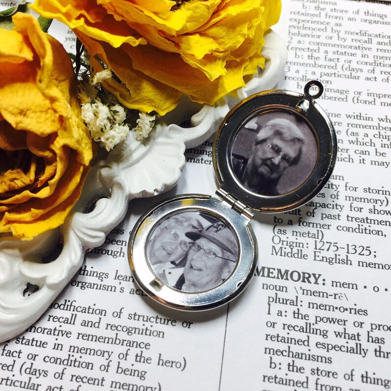 May include: A silver locket with a hinged lid, open to reveal a black and white photo of two people. The locket is lying on a page of a book with the word "MEMORY" printed in large letters.