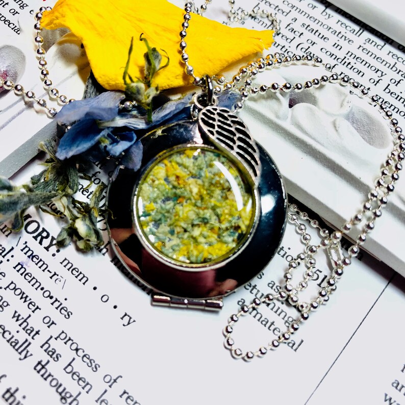 May include: A silver locket necklace with a small, round glass window containing dried flowers. The locket has a silver wing charm attached to it. The necklace is on a white surface with text that reads "MEMORY: mem...ory".