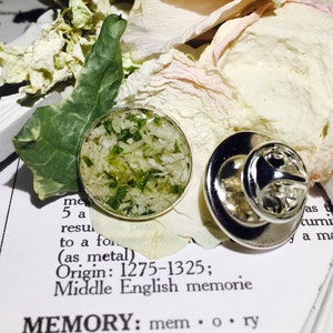 May include: A silver pin with a round, clear resin top containing pressed green and white botanicals. The pin is on a white surface with a green leaf and a white flower petal.