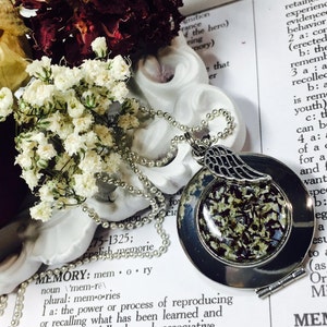 May include: A silver locket necklace with a small angel wing charm. The locket is open and filled with white and black dried flowers. The necklace is laying on a white surface with a dictionary page visible in the background. The page is open to the definition of the word "memory".