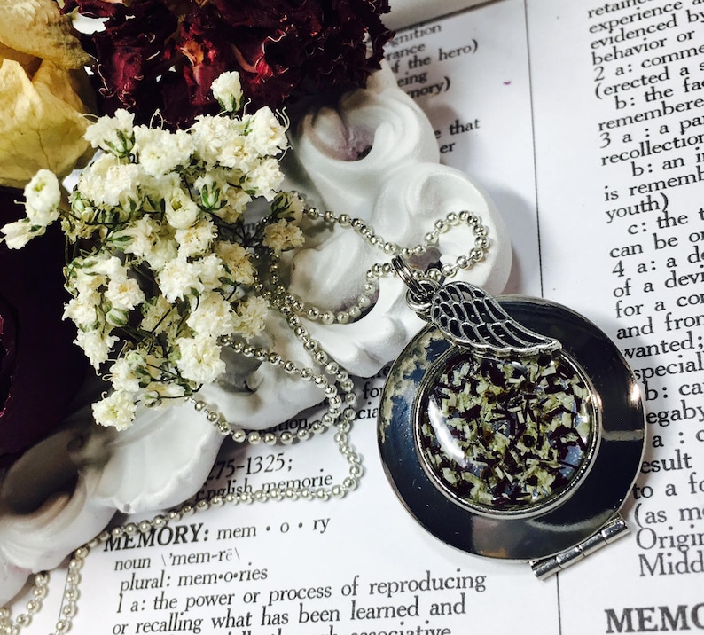 May include: A silver locket necklace with a small angel wing charm. The locket is open and filled with white and black dried flowers. The necklace is laying on a white surface with a dictionary page visible in the background. The page is open to the definition of the word "memory".