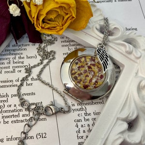 May include: A silver locket necklace with a small, round, clear glass window. The window contains dried flower petals in shades of red and yellow. The locket has a silver wing charm attached to it.