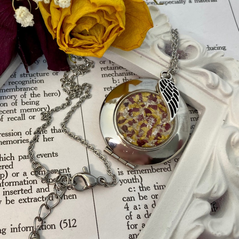 May include: A silver locket necklace with a small, round, clear glass window. The window contains dried flower petals in shades of red and yellow. The locket has a silver wing charm attached to it.