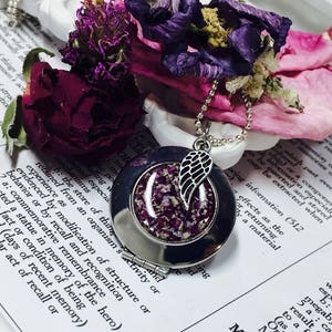 May include: A silver locket necklace with a small angel wing charm. The locket is open, revealing a clear resin filled with dried purple and pink flower petals.