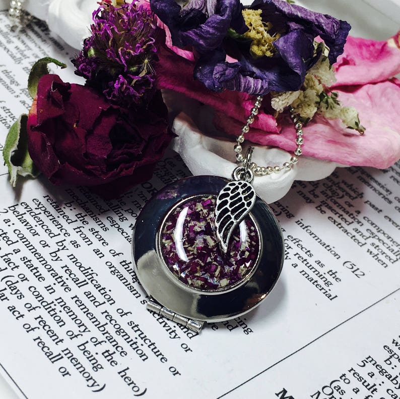 May include: A silver locket necklace with a small angel wing charm. The locket is open, revealing a clear resin filled with dried purple and pink flower petals.