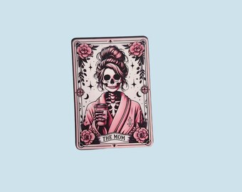 May include: A tarot card with a pink and black illustration of a skeleton woman wearing a pink robe and holding a cup of coffee. The card is titled "The Mom".