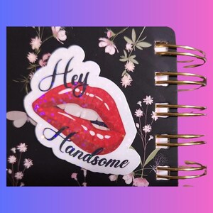 May include: A black spiral notebook with a white sticker that says "Hey Handsome" and features a red glitter lip print.