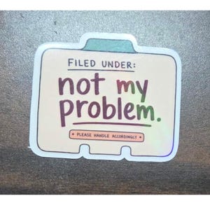 May include: A rectangular sticker shaped like a file folder with a cream-coloured background. It features the text FILED UNDER: not my problem. in dark purple and green, with a smaller box below containing the text PLEASE HANDLE ACCORDINGLY.