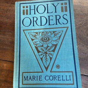 May include: A blue book with the title "HOLY ORDERS" and the author "MARIE CORELLI" on the cover. The cover features a gold triangle with a stylized flower design.