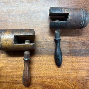 May include: Two antique wooden clamps with screw mechanisms and wooden handles. The clamps are made of dark wood with visible wear and tear, suggesting age and use. The handles are a darker color than the clamp bodies.
