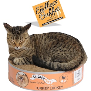 May include: A brown tabby cat resting on a round cardboard scratch pad designed to look like a large can of Chicken Puss Turkey Lurkey gourmet cat food. The packaging features a label with text, a cat image, and a net weight of 3 oz.