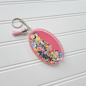 May include: Oval-shaped pink keychain with a silver chain and a pink tassel. The keychain contains colorful miniature candies and the text "I Spy". A fun accessory for keys or bags.