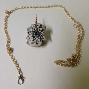 May include: A handmade necklace featuring a pendant with a black floral design on a cream-colored base. The pendant is suspended from a gold-colored chain with a lobster clasp. The pendant is approximately 1 inch in length.