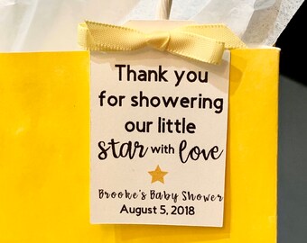 May include: A white card with black text that says "Thank you for showering our little star with love" and "Brooke's Baby Shower August 5, 2018". A gold star is above the text "love". The card is tied with a yellow ribbon and attached to a yellow gift bag.