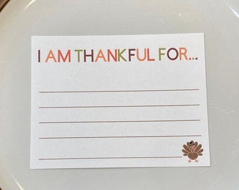 May include: A brown plate with a white rim and a white card on top. The card has the text "I AM THANKFUL FOR..." and a small turkey illustration. The plate is on a woven placemat.