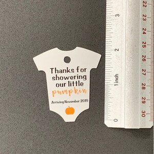 K&ouml;nnte beinhalten: Wei&szlig;er Baby-Strampler-f&ouml;rmiger Dankesanh&auml;nger mit dem Text "Danke, dass du unser kleines K&uuml;rbischen geduscht hast Ankunft im November 2019" und einer kleinen orangefarbenen K&uuml;rbisgrafik.