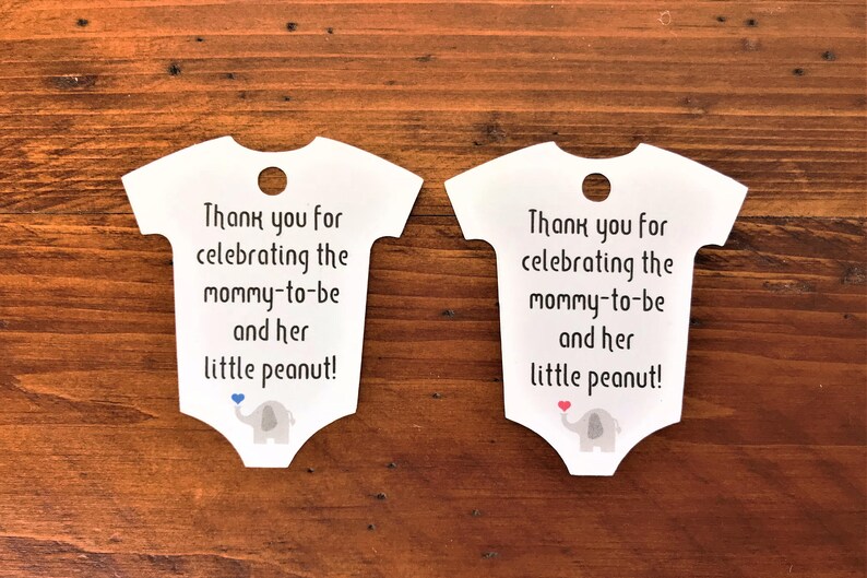 May include: Two white baby onesie shaped thank you tags with a gray elephant holding a heart. The text on the tags reads "Thank you for celebrating the mommy-to-be and her little peanut!"