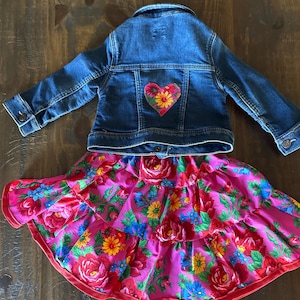 May include: A denim jacket with a heart-shaped floral patch on the back, paired with a tiered skirt. The skirt features a vibrant floral pattern in shades of pink, red, yellow, and blue. The outfit is designed for children.
