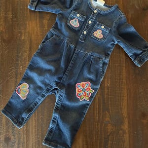 May include: A dark blue denim jumpsuit for a baby or toddler. The jumpsuit has long sleeves, pockets, and decorative floral and butterfly patches. It features a button-up front and is made of a soft denim material.