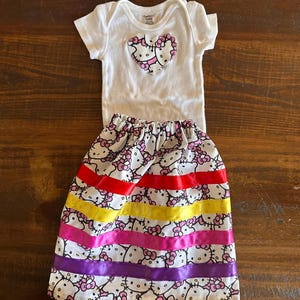 May include: A white short-sleeved bodysuit featuring a heart-shaped Hello Kitty applique on the chest, paired with a matching white skirt patterned with Hello Kitty faces and accented by horizontal red, yellow, pink, and purple satin ribbons.