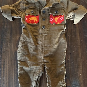 May include: Brown baby jumpsuit with a collared neckline and button-down front. The chest pockets feature colorful patterned fabric with a bison and bull design. The sleeves are rolled up, and the jumpsuit has a relaxed fit.