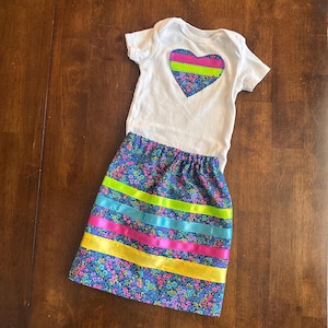 May include: A white baby bodysuit with a heart-shaped design in the center, featuring a floral pattern and neon green and pink stripes. The outfit includes a matching skirt with a floral pattern and colorful ribbon accents.