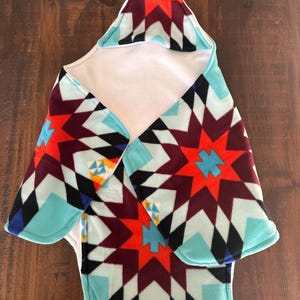 May include: A baby blanket with a geometric pattern in shades of red, turquoise, black, and white. The blanket has a hood and a soft, plush texture. The design features star-like shapes and diamond patterns.