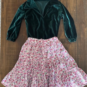 May include: A green velvet long-sleeved shirt with a v-neck collar and a white skirt with a pink and purple floral print.