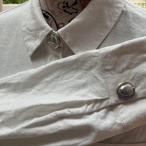Puede incluir: Una camisa blanca de manga larga con cuello clásico y puños abotonados. La camisa está adornada con distintivos botones de tono plateado, cada uno con forma de bellota texturizada, añadiendo un detalle metálico en el cuello y el puño.