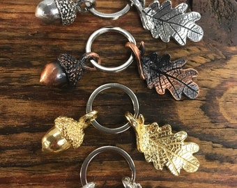 May include: Four metal keychains featuring acorn and oak leaf charms. The keychains have silver, gold, and copper finishes. The acorns have textured caps, and the oak leaves have detailed veining. The keyrings are silver.
