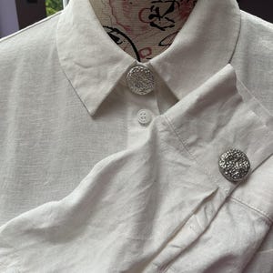 Peut inclure: Une chemise blanche à manches longues avec un col classique et une fermeture boutonnée. La chemise est agrémentée de gros boutons circulaires argentés à la surface texturée et martelée sur le col et les poignets des manches.