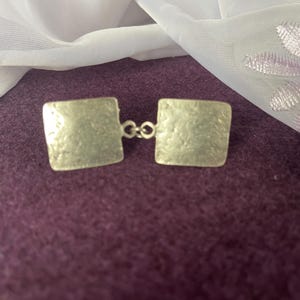 May include: A pair of silver square cufflinks with a textured surface and a connecting chain.