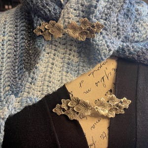 Puede incluir: Dos broches de hoja plateada se muestran en una bufanda de ganchillo azul y blanca. La bufanda est&aacute; drapeada sobre una camisa negra.