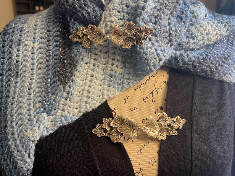 Puede incluir: Dos broches de hoja plateada se muestran en una bufanda de ganchillo azul y blanca. La bufanda est&aacute; drapeada sobre una camisa negra.