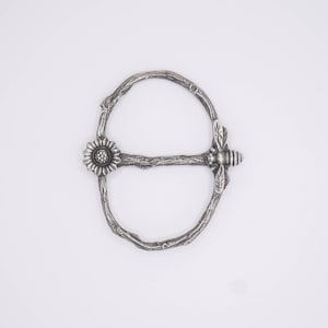 Pode incluir: Fivela de metal prateada com um design de galho, apresentando um girassol e uma abelha.