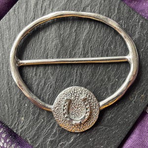 May include: A silver-toned belt buckle with a horseshoe design. The buckle has a circular frame with a horizontal bar and a textured, circular centerpiece featuring a horseshoe. The buckle is displayed on a dark, textured surface.