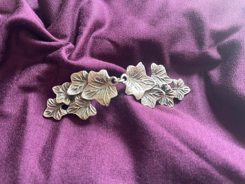 Puede incluir: Broche de metal plateado con dise&ntilde;o de hoja de hiedra. Las hojas est&aacute;n dispuestas en un racimo y tienen un aspecto detallado y realista.