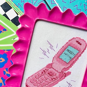 May include: A pink cross-stitch of a flip phone with the words "Anxiety Calling" on the screen, framed in a scalloped pink frame. The background includes colorful geometric patterns in teal, black, and pink.