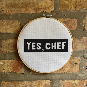 May include: A white embroidery hoop with a black rectangle that says "YES, CHEF" on a brick wall background.