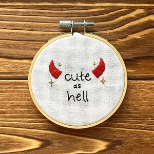 May include: A white embroidery hoop with a red devil horn design and the text "cute as hell".