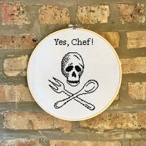 May include: A white embroidery hoop with the text "Yes, Chef!" and a black and white skull and crossbones design. The skull is wearing a chef's hat.