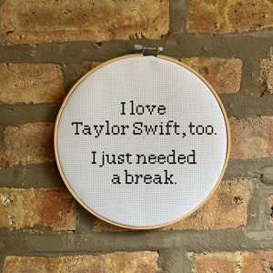 May include: A cross-stitch embroidery hoop with the text "I love Taylor Swift, too. I just needed a break." The text is in black on a white background. The hoop is set against a brick wall.