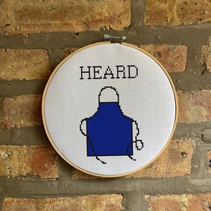 May include: A white embroidery hoop with a blue apron design and the word "HEARD" stitched in black thread.